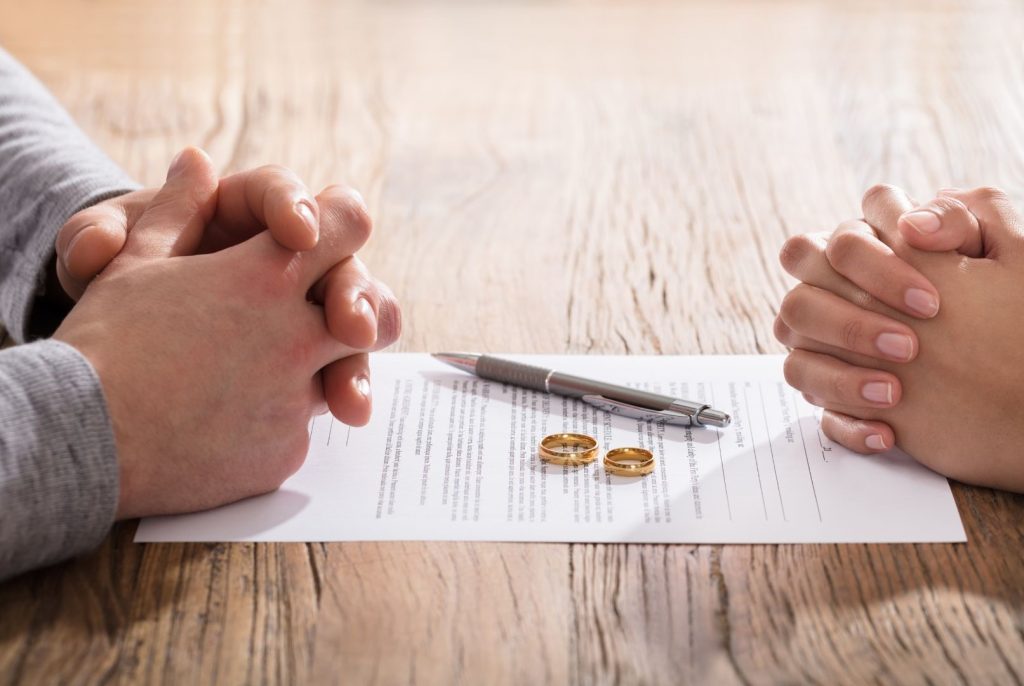 Two sets of clasped hands, with two marriage rings, a pen, and a form between them.