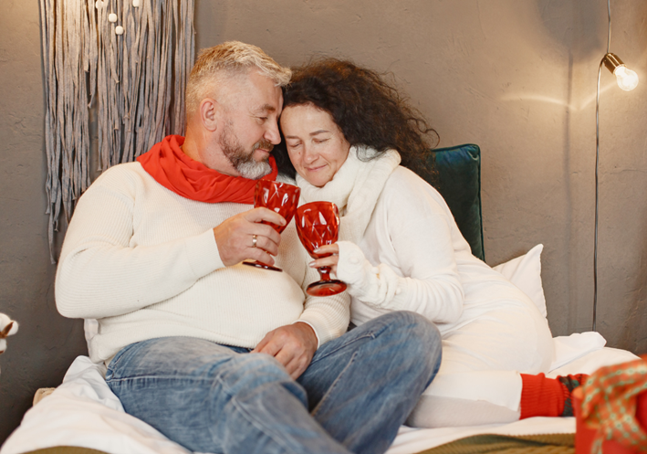 A middle-aged couple tapping drinks while lying in bed together.