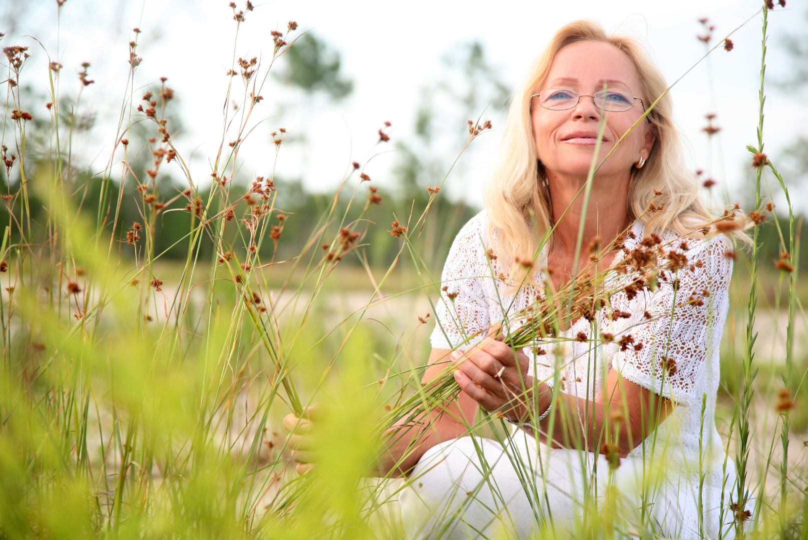 A middle-aged woman in nature, smiling as she crouches down in front of tall grass.