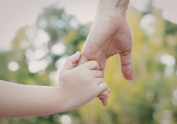A child's hand holding onto an adult's. The background is out of focus but appears to be of bright trees and an overcast day.