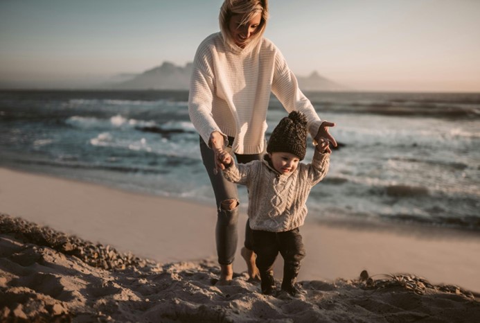 A mother and toddler on a beach. The mother is helping the toddler walk across the sand. They are both smiling.
