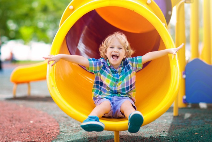 A blonde-haired boy coming out of a yellow tunnel slide, smiling widely.