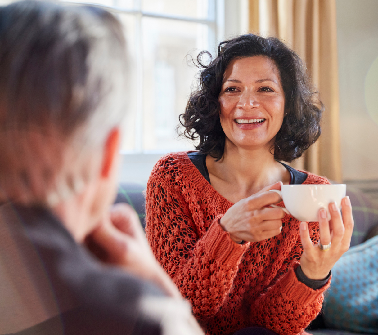 A woman in a russet jumper smiling whilst enjoying a hot drink.