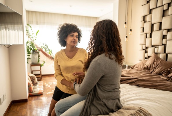A lesbian couple sitting on their bed, speaking whilst holding hands.