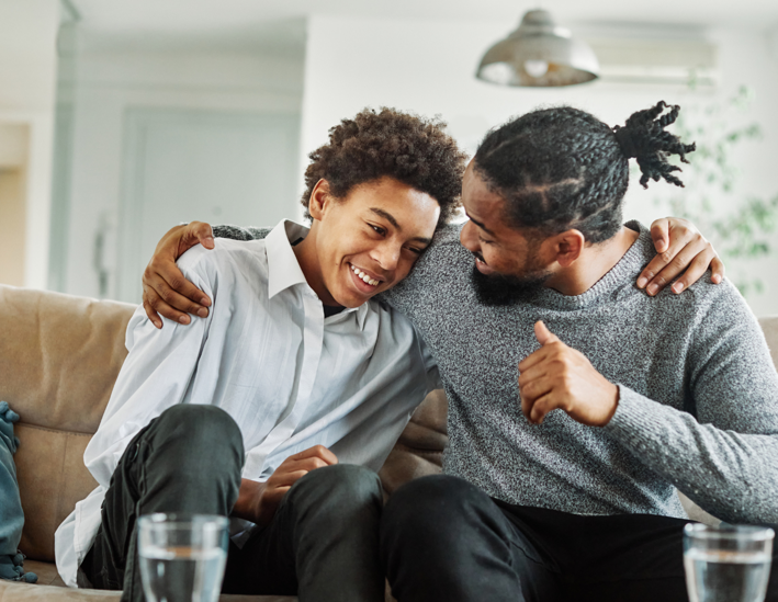 A dad and son with arms around each other, grinning while talking on the couch.