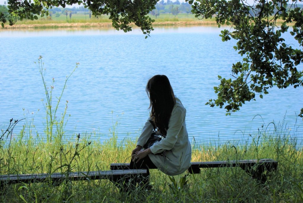A woman sitting on a bench by a countryside lake, her back to the camera.