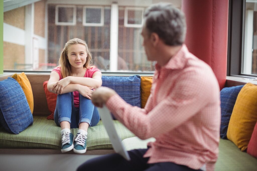 A teenage girl on a colourful sofa, talking to a male counsellor or Family Court Advisor. Her knees are drawn up, and her arms are resting on them.