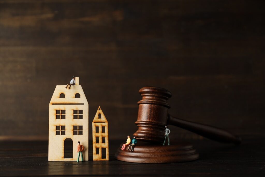 A 2D wooden model facade of two homes next to a gavel. Small plasticine people are sitting/standing around. The background is dark wood.